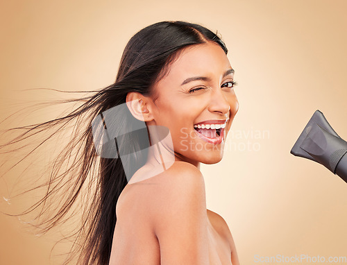 Image of Wink, hair and portrait of happy woman with hairdryer for beauty isolated in a studio brown background with strong texture. Aesthetic, glow and young person with heat or wind for health and shine