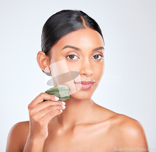 Image of Portrait, skincare and gua sha facial massage with a woman in studio on white background holding a stone. Face, beauty and natural with a confident young model using antiaging jade to relax her skin