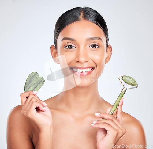 Image of Portrait, beauty and gua sha facial massage with a woman in studio on a white background holding a stone. Smile, skincare and face roller with a happy young model looking confident at luxury wellness