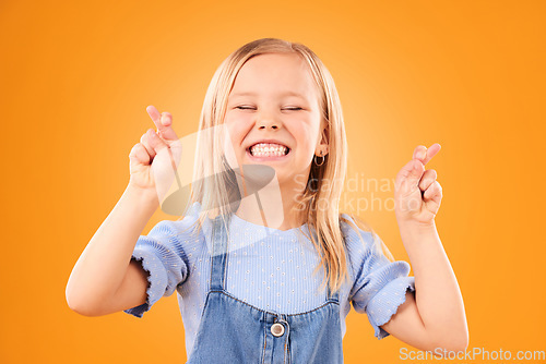 Image of Child, fingers crossed and wish or hope in studio for luck, prayer or faith. Face of excited young girl kid on a orange background for hand gesture, icon or sign for dream, optimistic or superstition