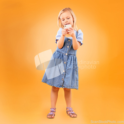 Image of Girl, child and typing with phone in studio for social media, play mobile games and download app on orange background. Young kid scroll smartphone to search website, watch multimedia and subscription