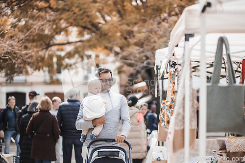 Image of Father walking carrying his infant baby boy child and pushing stroller in crowd of people wisiting sunday flea market in Malaga, Spain
