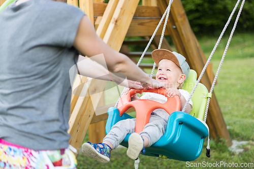 Image of Mother pushing her infant baby boy child on a swing on playground outdoors.