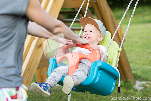 Image of Mother pushing her infant baby boy child on a swing on playground outdoors.