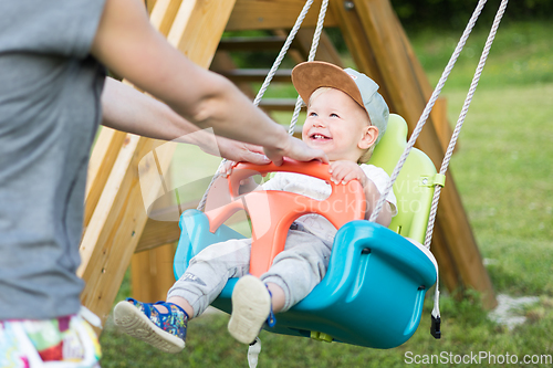 Image of Mother pushing her infant baby boy child on a swing on playground outdoors.