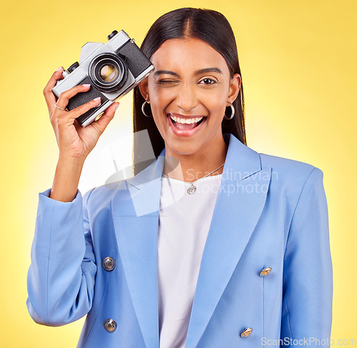 Image of Camera, photography and woman portrait with a smile and wink for picture in studio. Happy, female person and yellow background with creative, memory and funny emoji face with photographer skill
