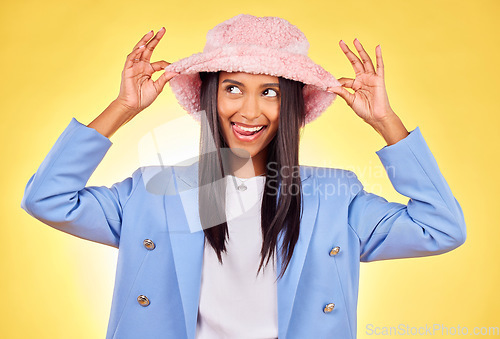 Image of Fashion, funny face and hat with a trendy indian woman on a yellow background in studio for style. Comic, smile and accessories with a happy young model sticking out her tongue in a cute outfit