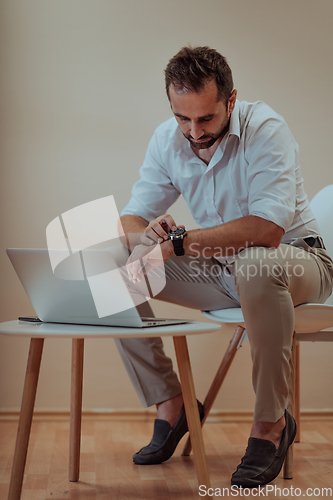 Image of A confident businessman sitting and using laptop and smartwatch with a determined expression, while a beige background enhances the professional atmosphere, showcasing his productivity and expertise.