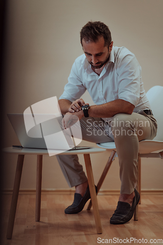 Image of A confident businessman sitting and using laptop and smartwatch with a determined expression, while a beige background enhances the professional atmosphere, showcasing his productivity and expertise.