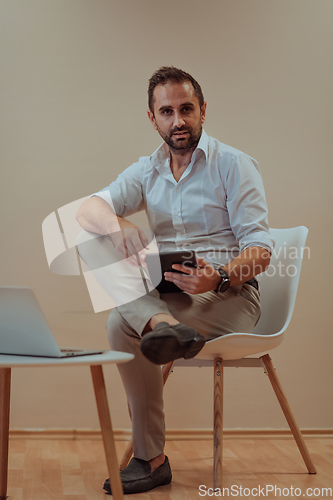 Image of A confident businessman sitting and using tablet with a determined expression, while a beige background enhances the professional atmosphere, showcasing his productivity and expertise.