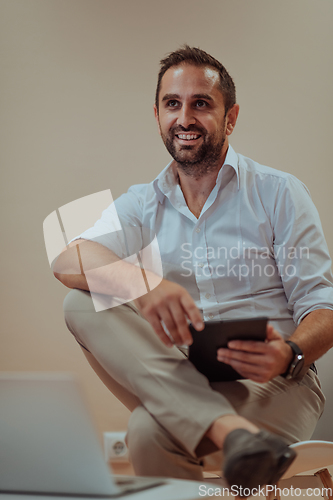 Image of A confident businessman sitting and using tablet with a determined expression, while a beige background enhances the professional atmosphere, showcasing his productivity and expertise.