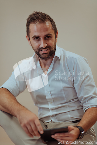 Image of A confident businessman sitting and using tablet with a determined expression, while a beige background enhances the professional atmosphere, showcasing his productivity and expertise.