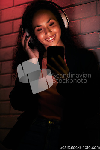 Image of Woman, phone and headphones for city or night streaming, listening to club music or party in neon light. Happy young person on mobile, social media and audio for techno, rave or network on brick wall