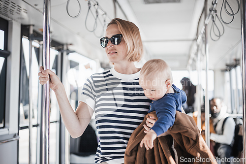 Image of Mother carries her child while standing and holding on to bar holder on bus. Mom holding infant baby boy in her arms while riding in public transportation. Cute toddler traveling with mother.