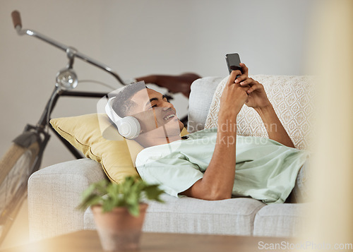 Image of Headphones, phone video and man on a living room sofa happy and relax at home. Series, funny app and laughing with music and listening in a house lounge with web mobile streaming and couch with smile