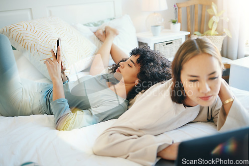 Image of Lesbian, couple and bed with laptop and phone for streaming movie or internet for relax, connection or online. Lgbt woman, reading and lens flare home for social media or subscription in bedroom