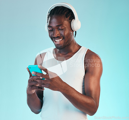 Image of Headphones, phone and black man in a studio networking and listening to music, playlist or album. Happy, smile and African male model scroll on internet with cellphone and song by blue background.