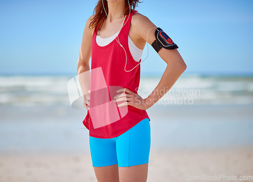 Image of Runner woman, beach fitness and music with earphones for podcast, workout and training motivation at ocean. Athlete girl, exercise or running for wellness by sea with focus, hands on hips or health