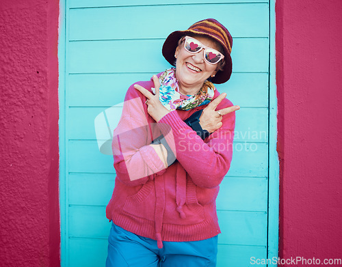 Image of Portrait, peace and funky with a senior woman outdoor standing against a blue door and red wall background. Glasses, hands and hip hop with a happy mature female doing a hand sign or gesture outside