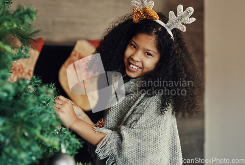 Image of Christmas, tree and decorating with a girl in antlers putting an ornament on a branch in the living room of her home. Children, portrait and smile with a happy kid placing decorations for celebration