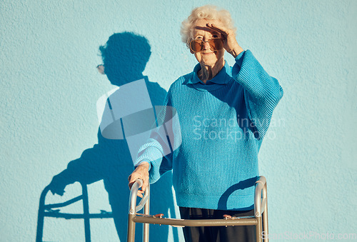 Image of Portrait, shadow and disability with a senior woman on a blue wall background while holding a mobility walker outdoor. Health, handicap and fashion with a mature female standing outside alone