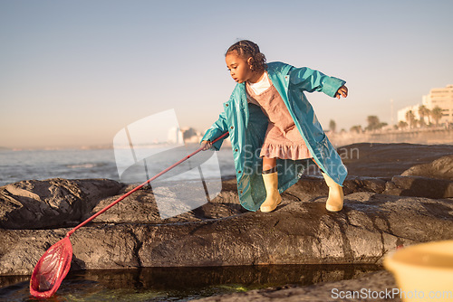 Image of Children, beach and fishing with a girl using a net in a rock pool while having fun on summer vacation. Water, nature and kids with a black child outdoor by a tide pool to fish and enjoy her holiday