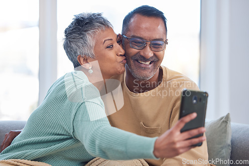 Image of Phone, selfie and kiss with a senior couple posing for a picture in the living room of their home together. Smartphone, kissing and social media with a mature man and woman taking a photograph