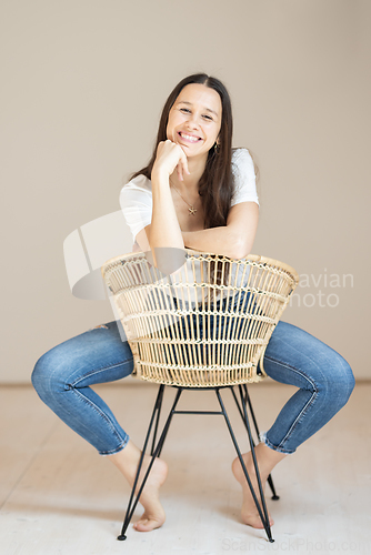 Image of Portrait of confident beautiful woman with long brown hair, wearing casual clothes, sitting on chair in tight jeans and white t-shirt, studio background