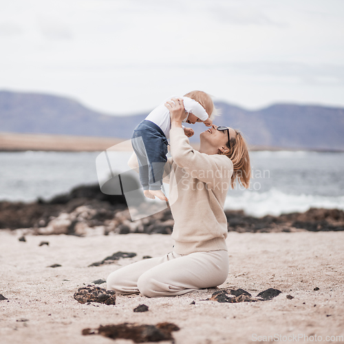 Image of Mother enjoying winter vacations holding, playing and lifting his infant baby boy son high in the air on sandy beach on Lanzarote island, Spain. Family travel and vacations concept