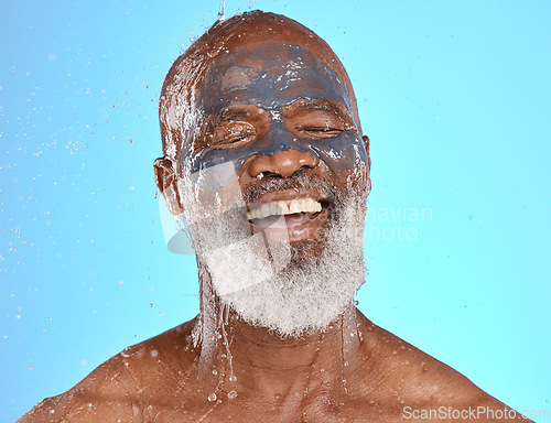 Image of Face, water splash and senior black man in clay mask in studio isolated on a blue background. Cleaning, retired and elderly male from Nigeria washing off facial cosmetics for skincare or healthy skin