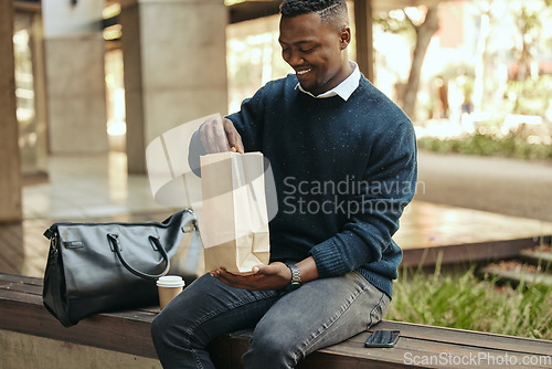 Image of Office employee with a coffee, a paper sandwich bag and a smile, on a bench for a break from his busy work schedule. Happy, hungry businessman, time to eat, with his food and drink outside for lunch.