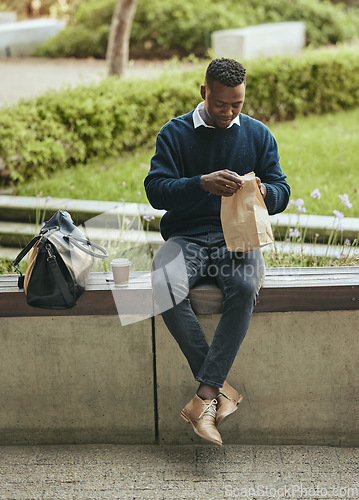 Image of Business man eating food for lunch on his break outdoors at a park opening a brown paper bag. Hungry, happy black male time to eat and drink outside in the city. Enjoying a takeaway or takeout meal