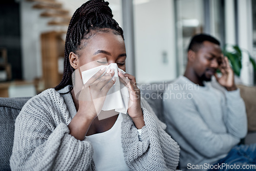 Image of Young, sick and ill woman blowing her nose with tissues while sitting on the couch at home. African lady has cold or flu and needs medicine. Husband worried about wife suffering from illness