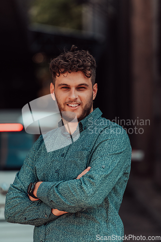 Image of A successful young businessman in a shirt, with crossed arms, poses outdoors, confident expression on his face.