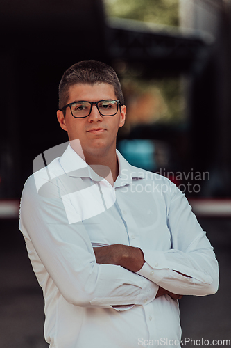 Image of A successful businessman in a white shirt, with crossed arms, poses outdoors, confident expression on his face.