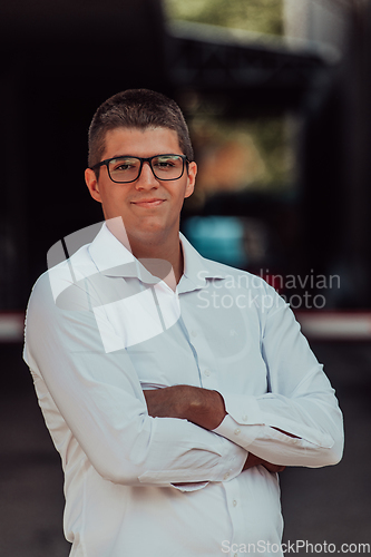 Image of A successful businessman in a white shirt, with crossed arms, poses outdoors, confident expression on his face.