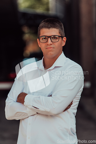 Image of A successful businessman in a white shirt, with crossed arms, poses outdoors, confident expression on his face.