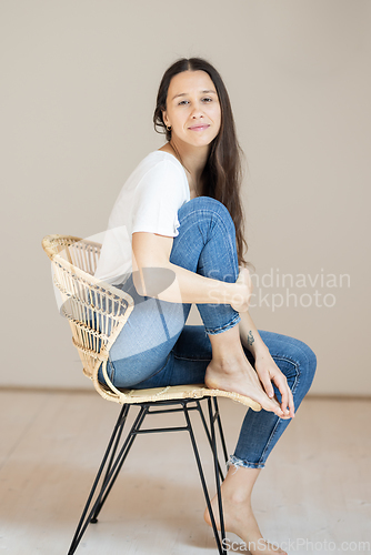 Image of Portrait of confident beautiful woman with long brown hair, wearing casual clothes, sitting on chair in tight jeans and white t-shirt, studio background