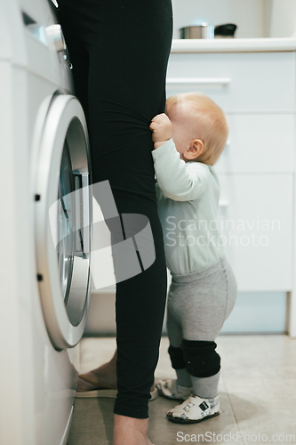 Image of Little infant baby boy child hiding between mothers legs demanding her attention while she is multitasking, trying to do some household chores in kitchen at home. Mother on maternity leave.