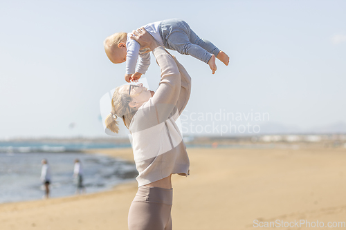 Image of Mother enjoying summer vacations holding, playing and lifting his infant baby boy son high in the air on sandy beach on Lanzarote island, Spain. Family travel and vacations concept.