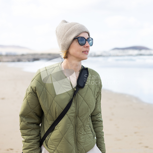 Image of Portrait of young stylish woman wearing green padded jacket, hoodie, wool cap and sunglasses on long sandy beach in spring.