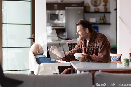 Image of Father wearing bathrope spoon feeding hir infant baby boy child sitting in high chair at the dining table in kitchen at home in the morning.