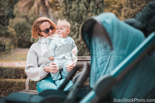 Image of Mother sitting on bench in urban park, laughing cheerfully, holding her smiling infant baby boy child in her lap having baby stroller parked by their site.