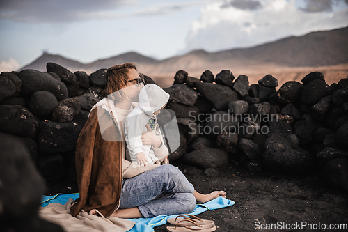 Image of Mother enjoying winter vacations playing with his infant baby boy son on black sandy volcanic beach of Janubio on Lanzarote island, Spain on windy overcast day. Family travel vacations concept.