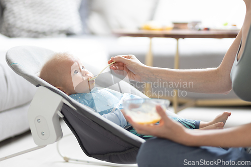 Image of Mother spoon feeding her baby boy infant child in baby chair with fruit puree. Baby solid food introduction concept.