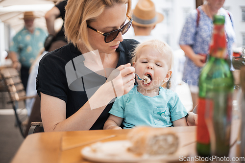 Image of Young caucasian blonde mother spoon feeding her little infant baby boy child outdoors on restaurant or cafe terrace in summer.