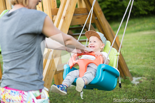 Image of Mother pushing her infant baby boy child on a swing on playground outdoors.