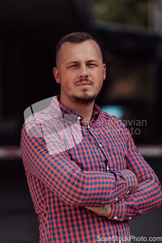 Image of A successful young businessman in a shirt, with crossed arms, poses outdoors, confident expression on his face.