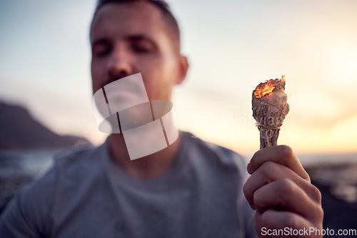 Image of Man, hand and burning sage on the beach while meditating for spiritual peace and zen mindfulness. Herb, burn and alternative cleanse ceremony to meditate for smudge during a seaside peaceful time