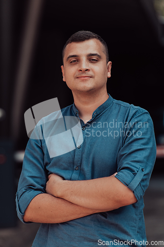 Image of A successful young businessman in a shirt, with crossed arms, poses outdoors, confident expression on his face.